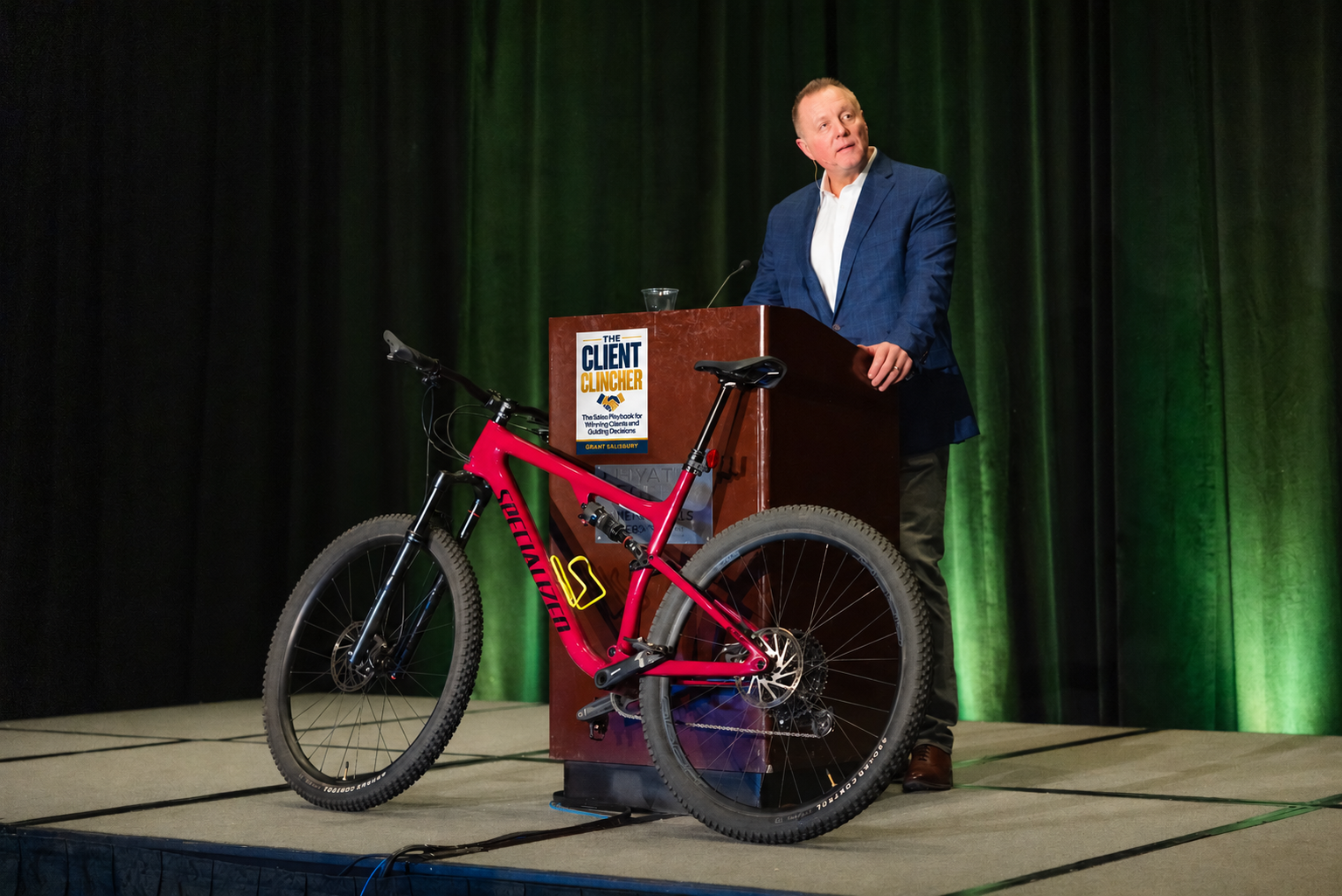 Grant Salisbury presenting beside a bicycle, with The Client Clincher book cover displayed on the podium.
